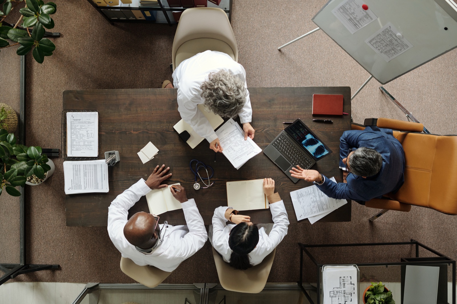 Overhead view of a physician team meeting with a medical billing consultant over patient charts and a laptop, the kind of hands-on partnership MMSM brings Michigan providers